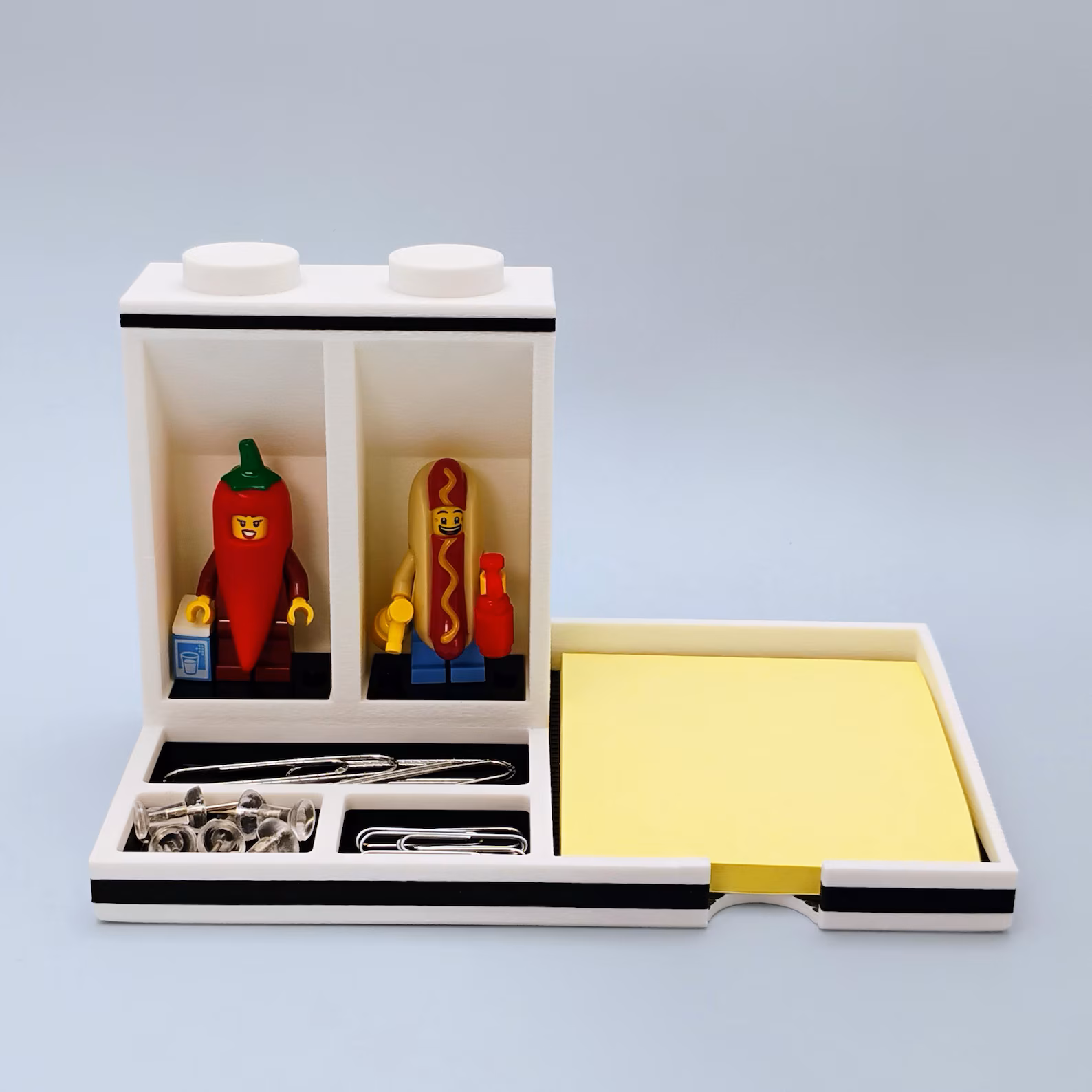 LEGO minifigure desk organizer with integrated Post-it note holder shown from the front. Features two minifigure display spots, an upright storage compartment, and a dedicated space for standard square sticky notes. Designed to keep desks organized while displaying favorite figures.